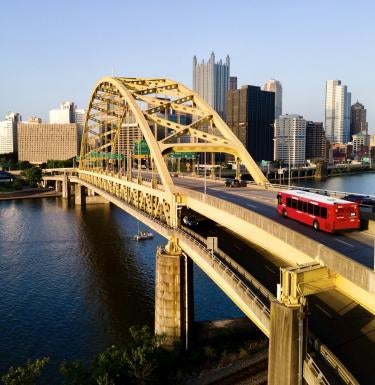 A red bus crosses a bridge with the city of Pittsburgh in the background 